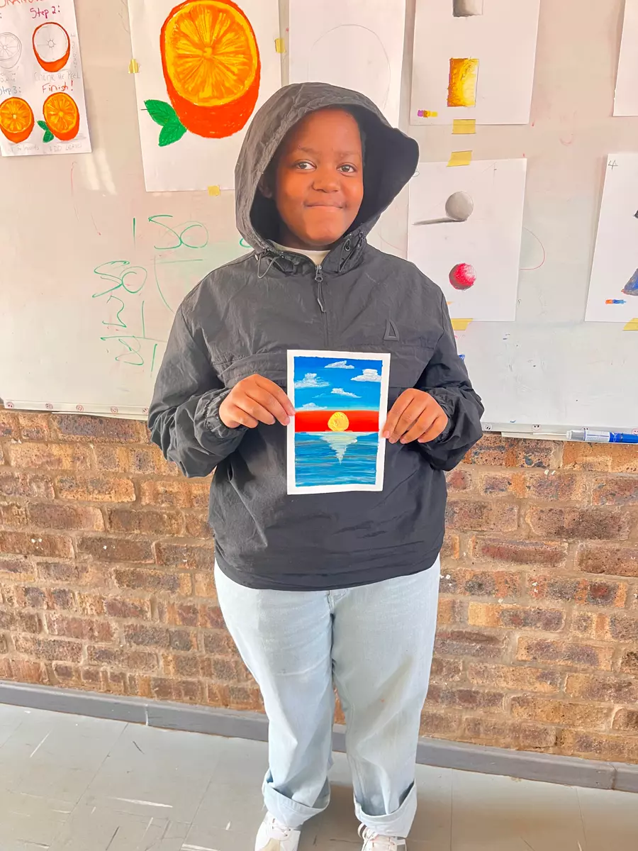 A child holding up a painting they made of a sunset over water