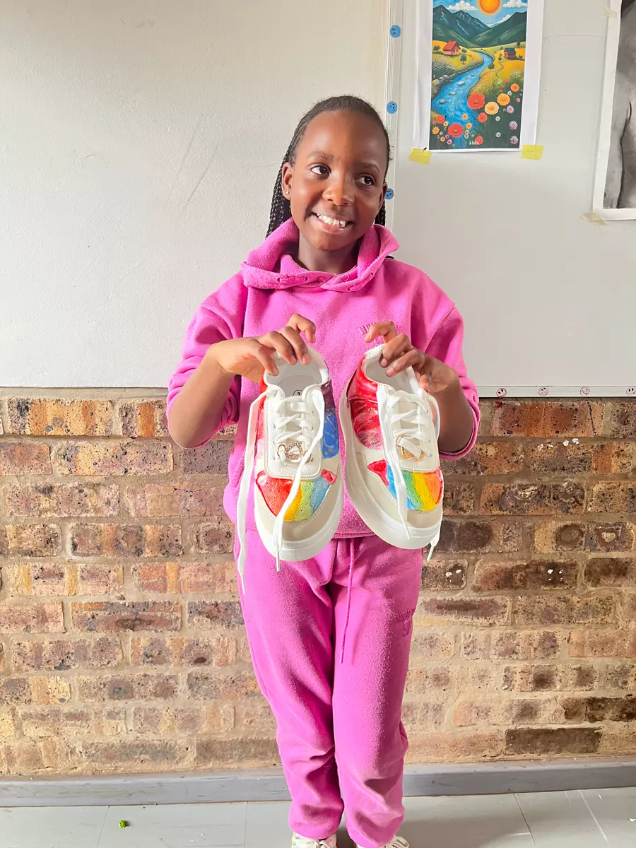 A young girl in a pink tracksuit holding up two painted white sneakers