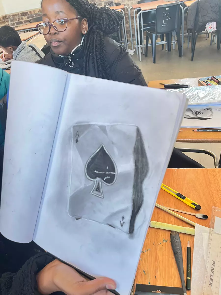 A student holding up an open sketchbook showing a pencil drawing of an ace of spades