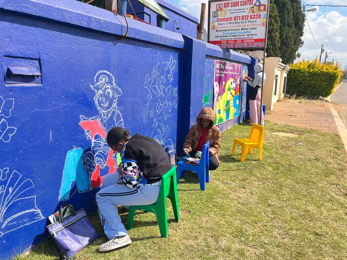 Three people painting a large blue wall with chalk and paint outside a day care centre
