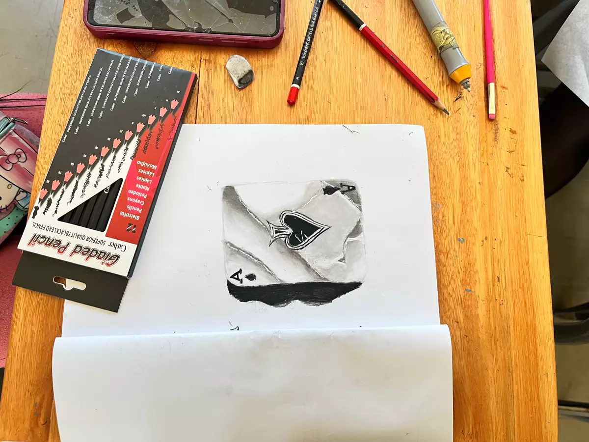 A pencil drawing of an ace of spades playing card on a wooden table with art supplies