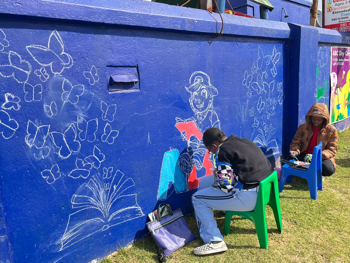 Two artists sitting on small chairs painting a long blue wall covered in chalk drawings